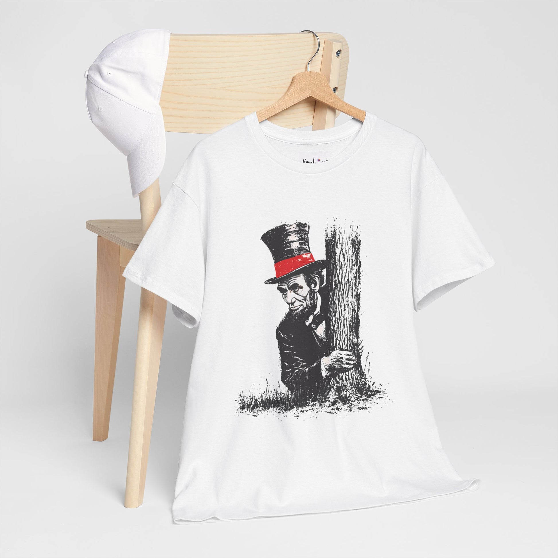 White t-shirt with a graphic design of a person in a top hat hiding behind a tree, hanging on a wooden hanger against a white background.